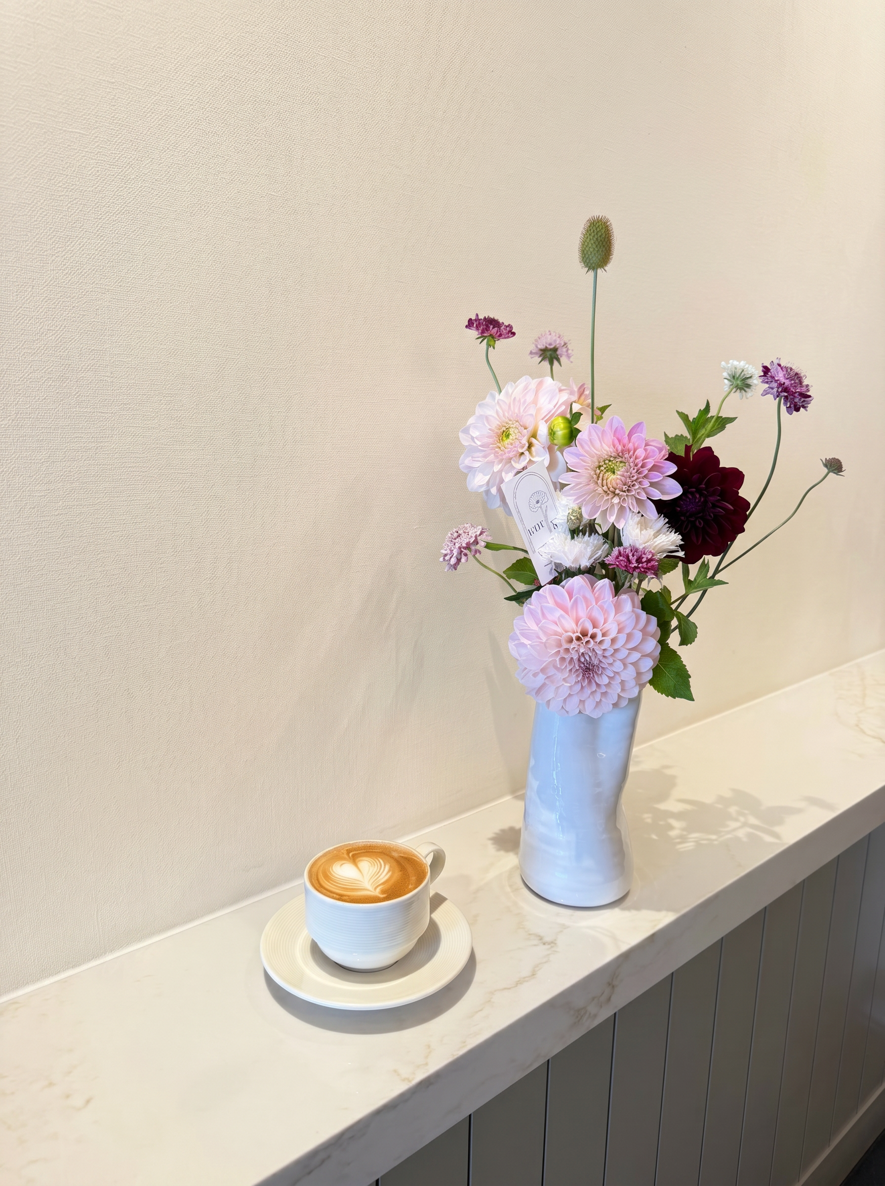 Pink dahlias and burgundy accents in a pale blue vase beside a latte