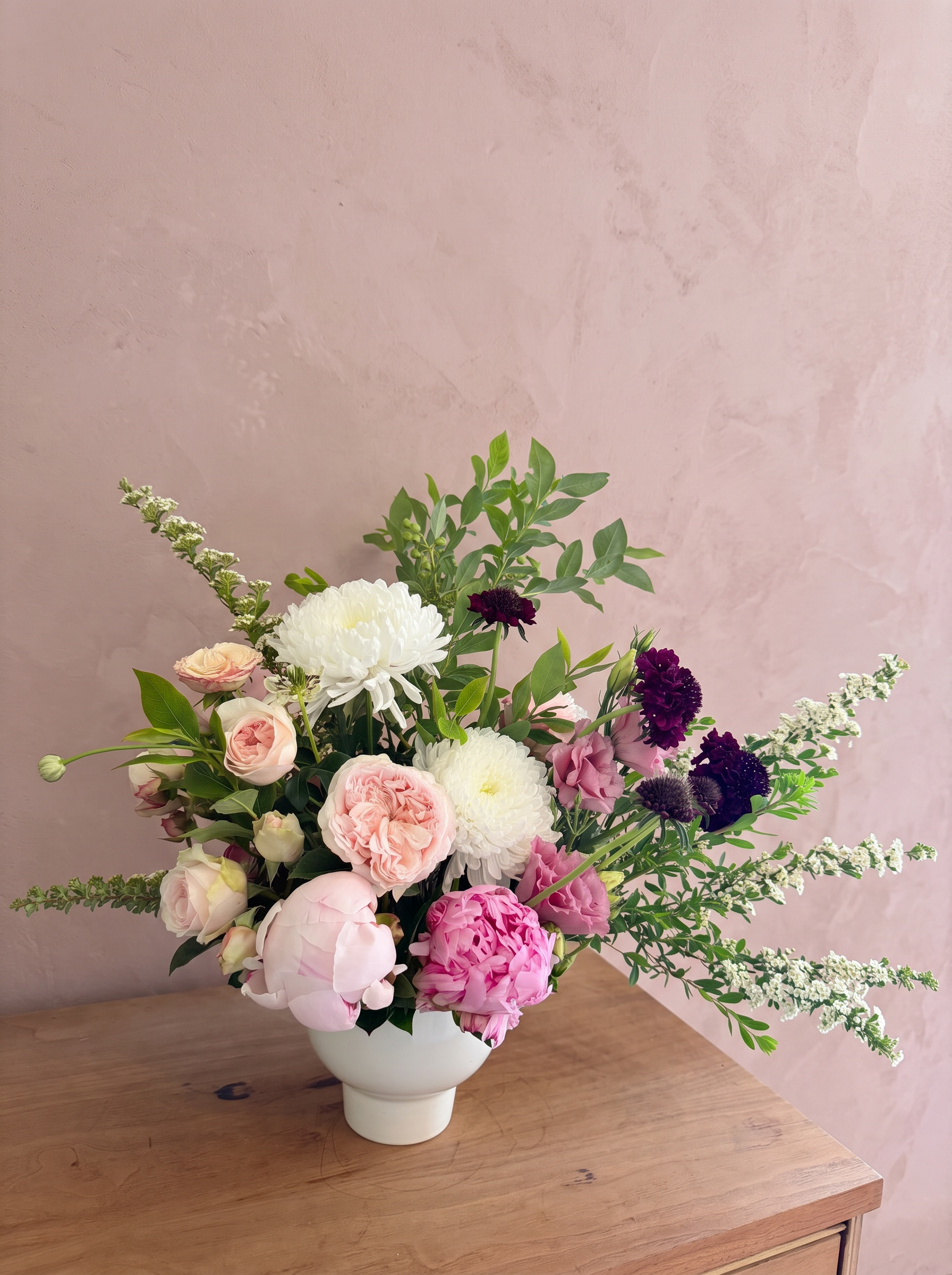 Lush pink peony and chrysanthemum arrangement with burgundy scabiosa in a white footed vessel
