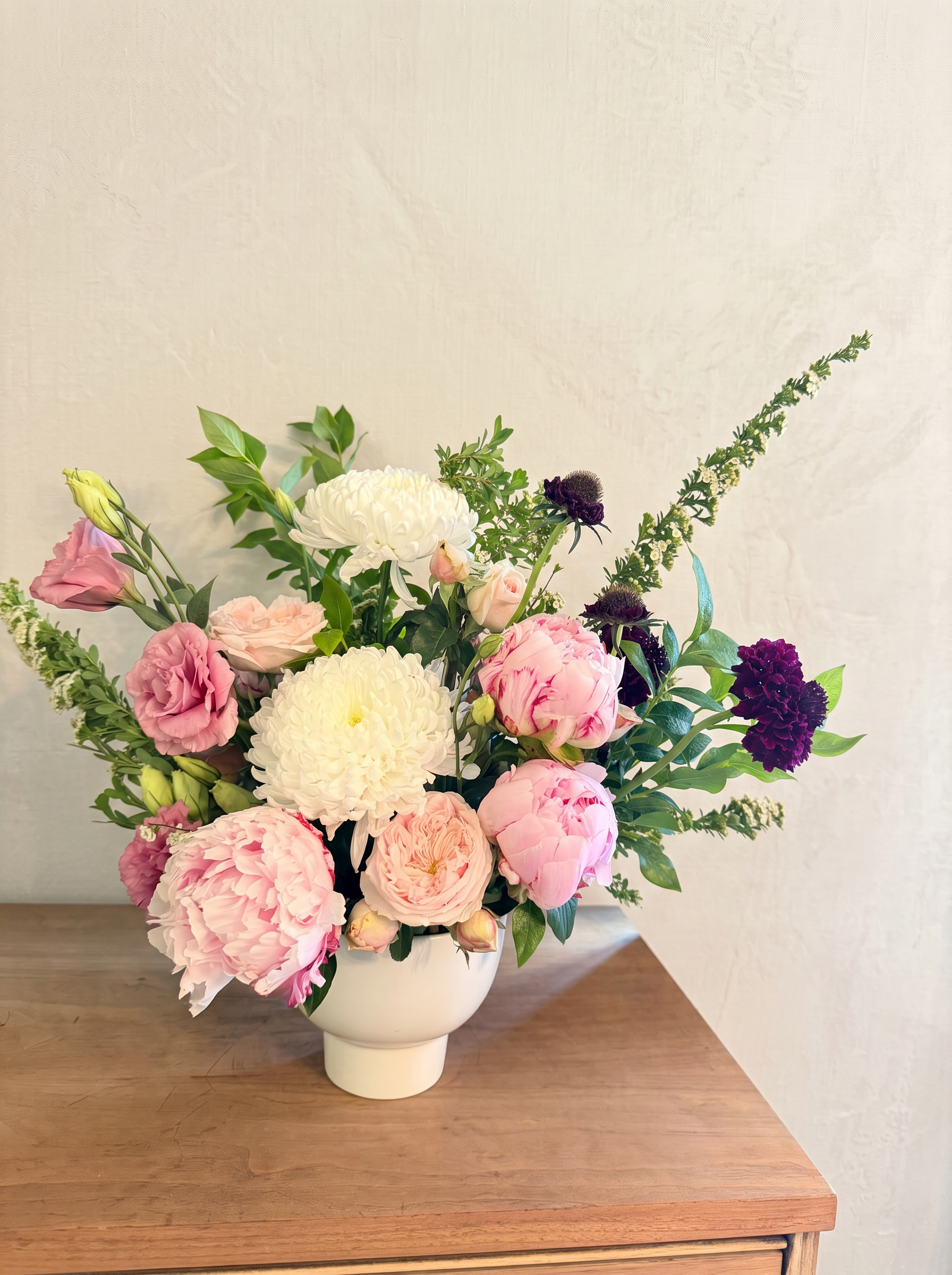 Pink peonies, white chrysanthemums and burgundy carnations in a footed white vessel