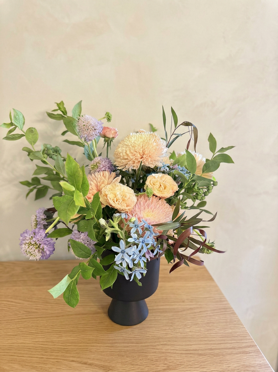 Peach chrysanthemum, lavender scabiosa and blue tweedia in a black footed vase on oak
